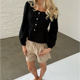 En kvinde med lyst, bølget hår står iført en sort bluse med knapper og beige shorts. Hun har sorte klipklappere på en grå beton- og hvid baggrund.