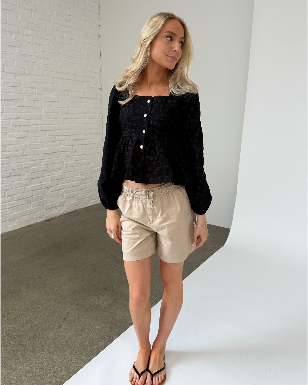 En kvinde iført sort bluse og beige shorts står afslappet, kigger til siden. Baggrunden er en væg af hvide mursten og et gråt gulv i et minimalistisk rum.
