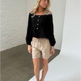 En kvinde iført sort bluse og beige shorts står afslappet, kigger til siden. Baggrunden er en væg af hvide mursten og et gråt gulv i et minimalistisk rum.
