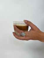 Hånden holder forsigtigt et glas med lagdelt kaffe og mælk. En dekorativ ring med en blomst er synlig på fingeren. Baggrunden er simpel og lys.
