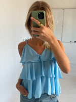 En person i en lyseblå, flæset top tager en selfie med en grøn telefon i et lyst rum med en tavle i baggrunden.