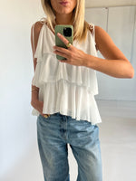 Kvinde iført hvid, lagdelt top og blå jeans tager spejlbillede med en grøn telefon. Hun står i et lyst rum, med én hånd i lommen og en gylden ring synlig.