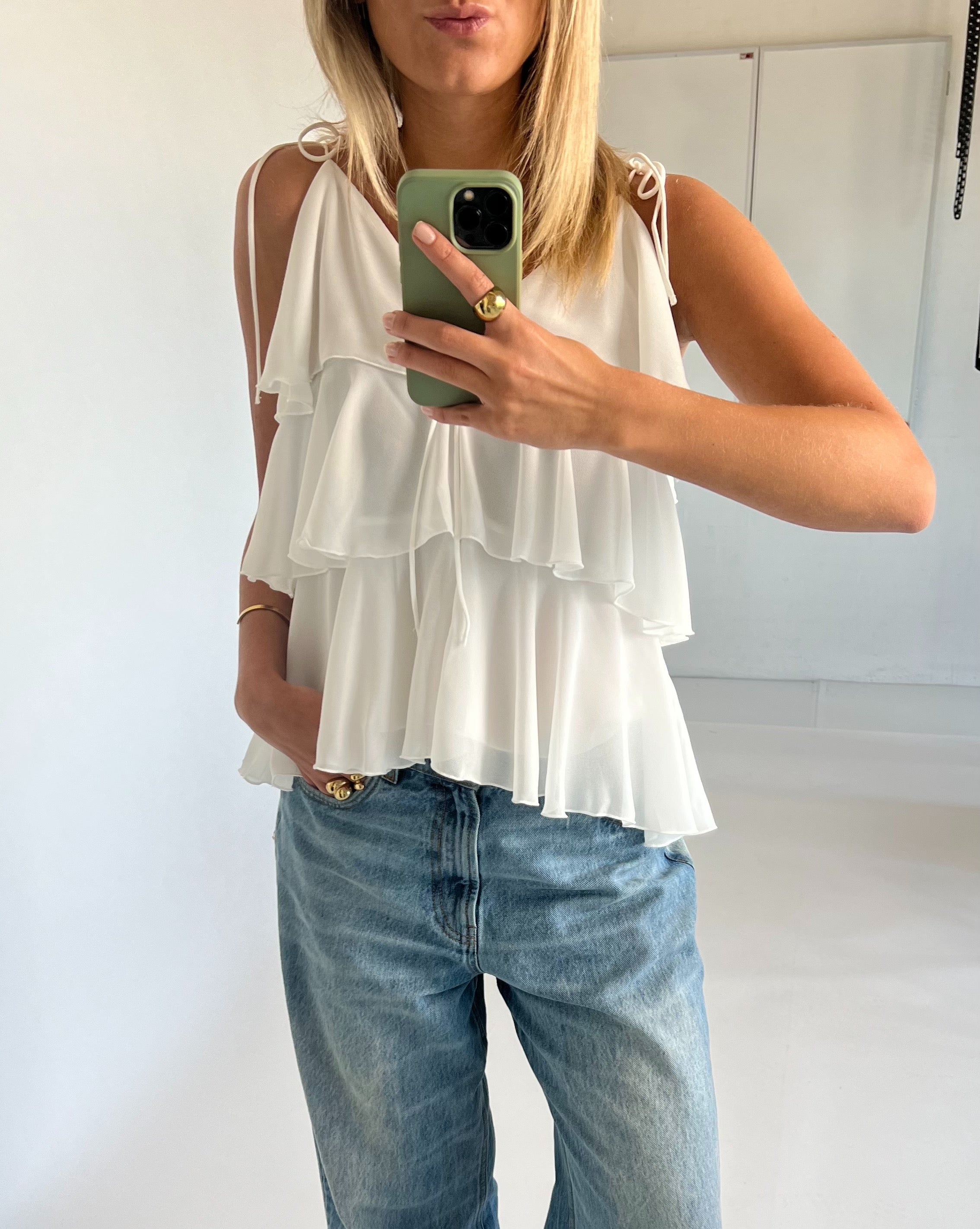 Kvinde iført hvid, lagdelt top og blå jeans tager spejlbillede med en grøn telefon. Hun står i et lyst rum, med én hånd i lommen og en gylden ring synlig.