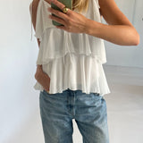 En person iført en hvid, lagdelt top tager et spejlbillede med en smartphone i et lysegråt rum. Personen har også løstsiddende jeans og en ring på fingeren.