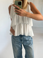 En person iført en hvid, lagdelt top tager et spejlbillede med en smartphone i et lysegråt rum. Personen har også løstsiddende jeans og en ring på fingeren.