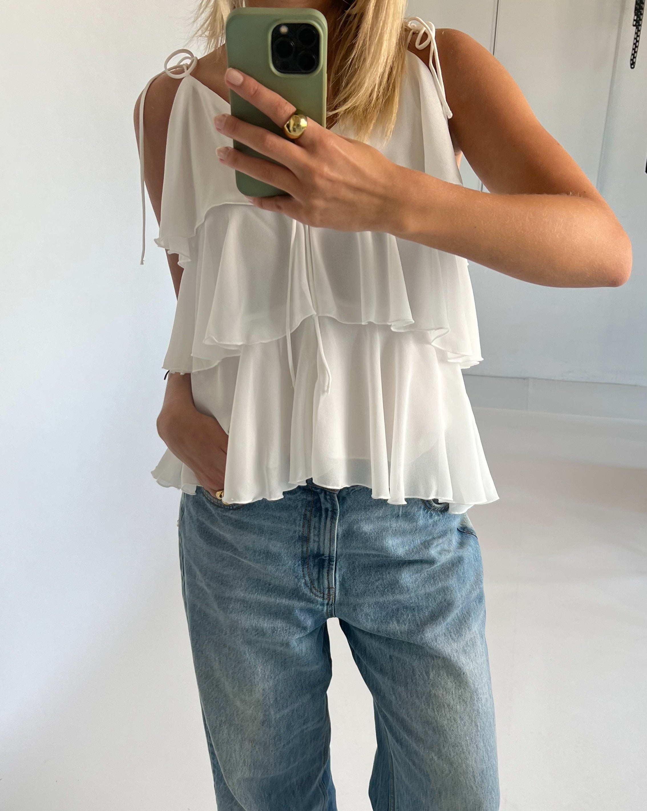 En person iført en hvid, lagdelt top tager et spejlbillede med en smartphone i et lysegråt rum. Personen har også løstsiddende jeans og en ring på fingeren.