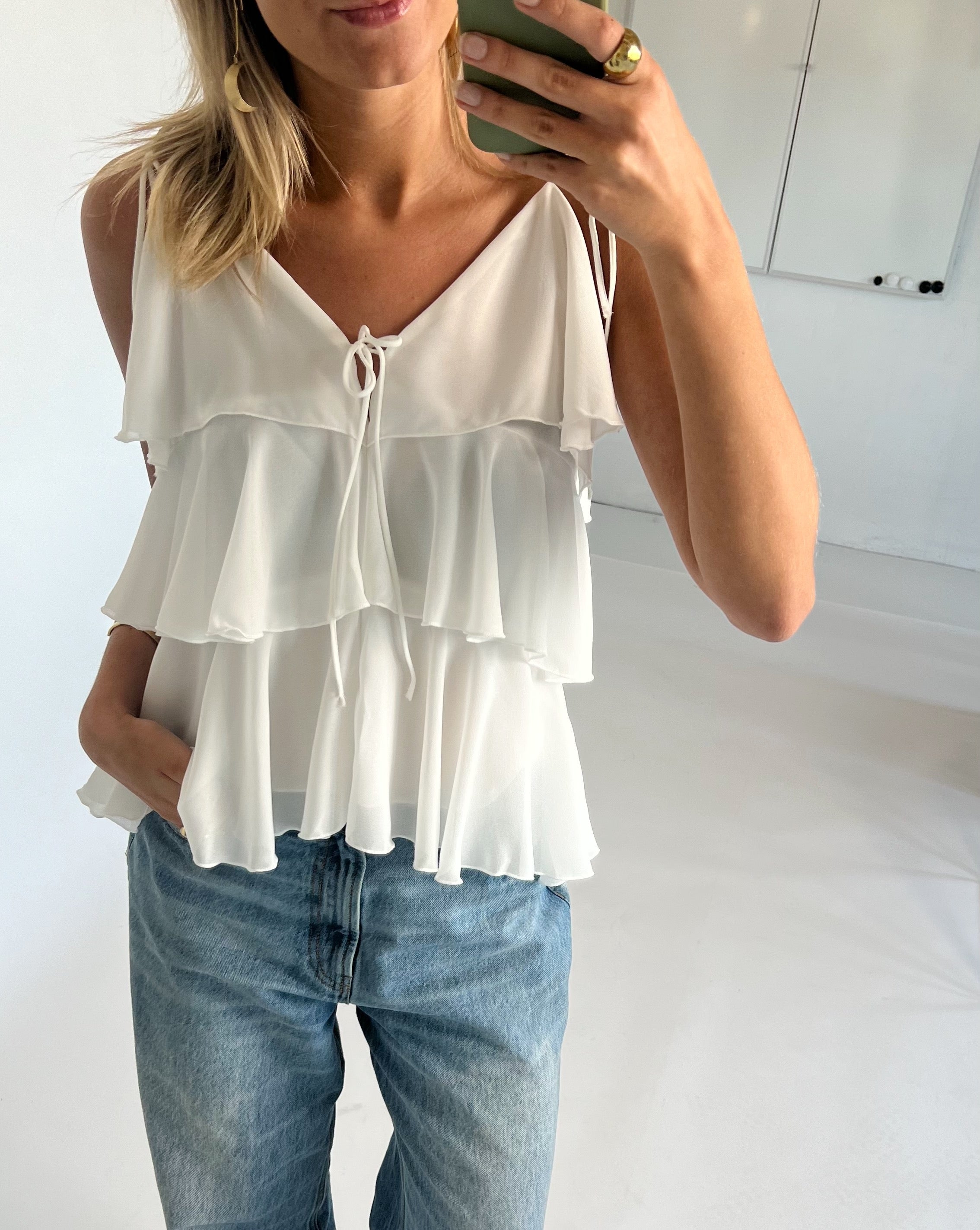 Kvinde iført hvid, flerlags top og blå jeans holder en smartphone, smiler og tager et spejlbillede. Håret er lys og skulderlangt, omgivet af et lyst interiør.