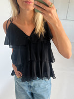 En person iført en sort, flæsebluse tager et spejlbillede, mens de holder en grøn telefon. De har også lyseblå jeans på. Rummet er lyst med hvide vægge.