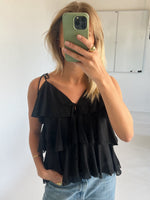 Kvinde står med en grøn telefon, dækker ansigtet, iført sort flæsetop og blå jeans, i et lyst rum med en hvid tavle i baggrunden.