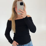 En person holder en telefon op foran ansigtet, iført en sort off-shoulder trøje og blå jeans, stående i et hvidt, neutralt rum.