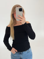 En person holder en telefon op foran ansigtet, iført en sort off-shoulder trøje og blå jeans, stående i et hvidt, neutralt rum.