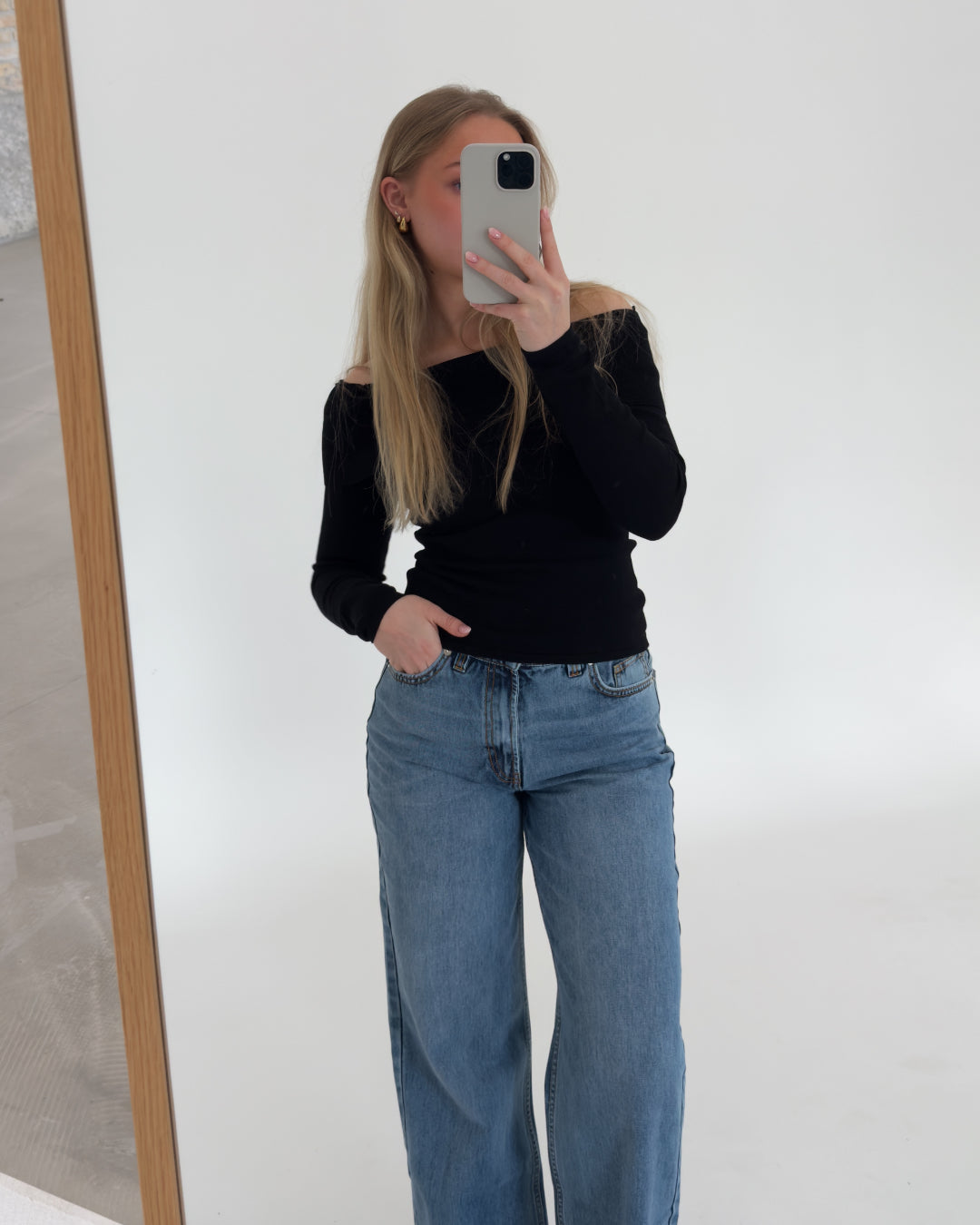 En person i sort trøje og blå jeans tager et spejlbillede med en telefon. Personen står i et lyst rum med en hvid væg og trægulv.