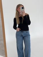 En person i sort trøje og blå jeans tager et spejlbillede med en telefon. Personen står i et lyst rum med en hvid væg og trægulv.