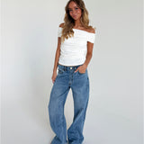 En person står afslappet iført en hvid top og blå jeans i et lyst, minimalistisk rum. Håret er langt og bølget, og hænderne er i lommen.