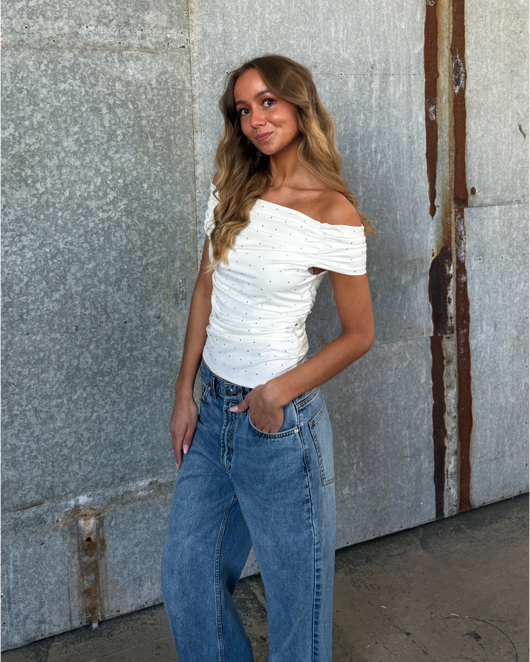 En ung kvinde står afslappet med hånden i lommen. Hun har en hvid off-shoulder-top og jeans på. Baggrunden er en rustik metalvæg.