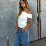 En ung kvinde står afslappet med hånden i lommen. Hun har en hvid off-shoulder-top og jeans på. Baggrunden er en rustik metalvæg.
