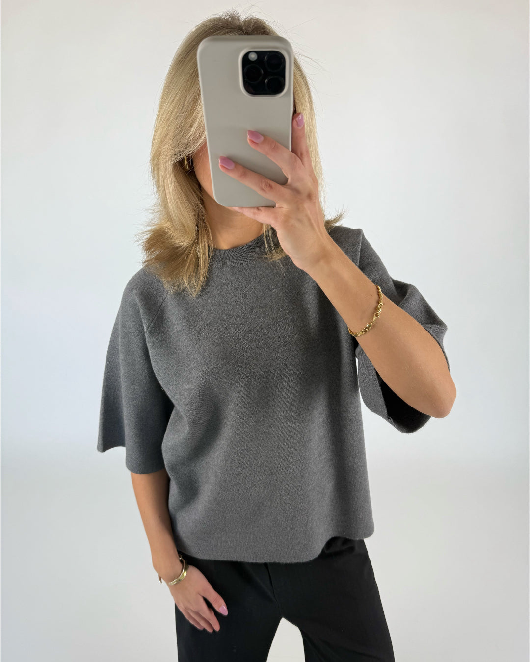 En person iført en grå trøje tager et spejlbillede med en smartphone i et neutralt, hvidt rum. Personen bærer også sort tøj og et guldfarvet armbånd.