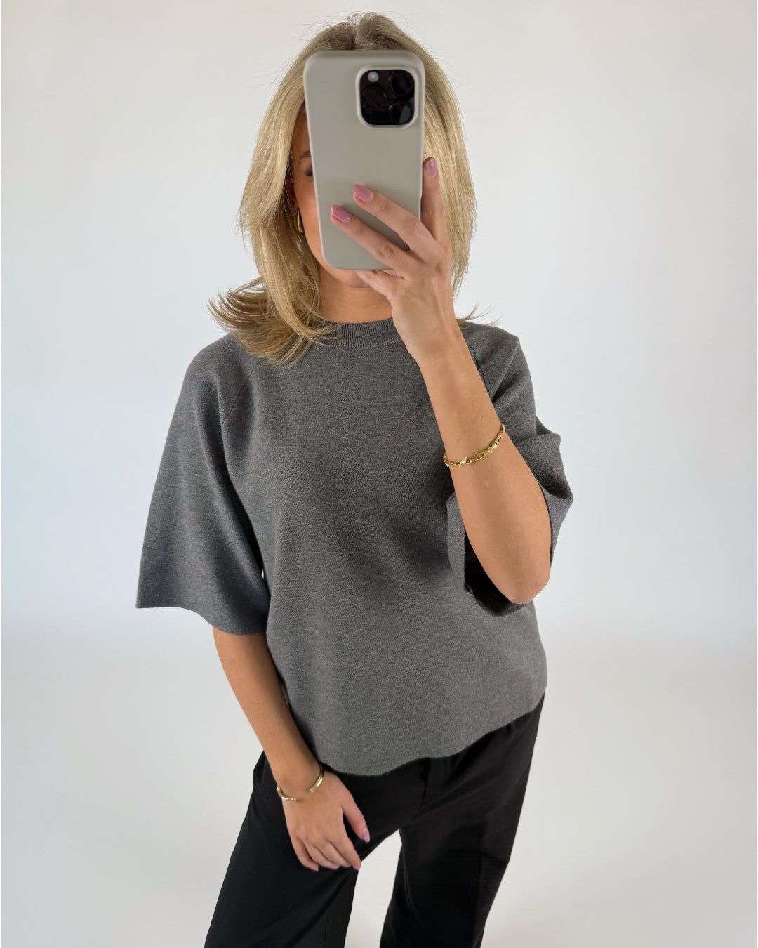 En person i gråt tøj tager et spejlbillede og holder en smartphone. Personen har blondt hår og et armbånd. Baggrunden er ensfarvet lys.