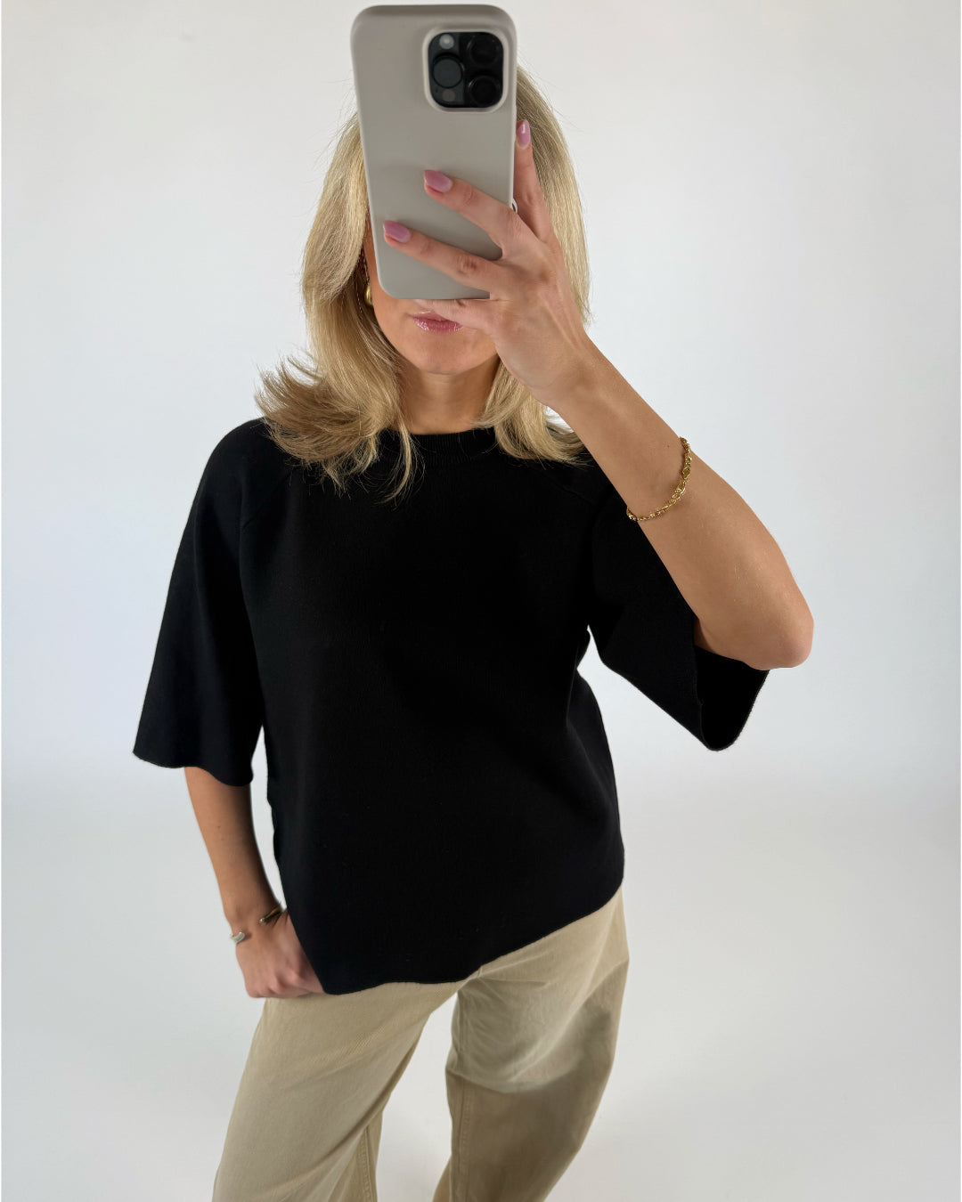 En person iført sort trøje og beige bukser tager et billede med en smartphone i et minimalistisk, lyst rum. De har en gylden armbånd på.