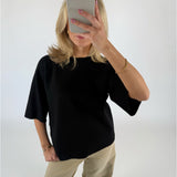 En person iført sort trøje og beige bukser tager et billede med en smartphone i et minimalistisk, lyst rum. De har en gylden armbånd på.