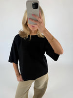 En person iført sort trøje og beige bukser tager et billede med en smartphone i et minimalistisk, lyst rum. De har en gylden armbånd på.