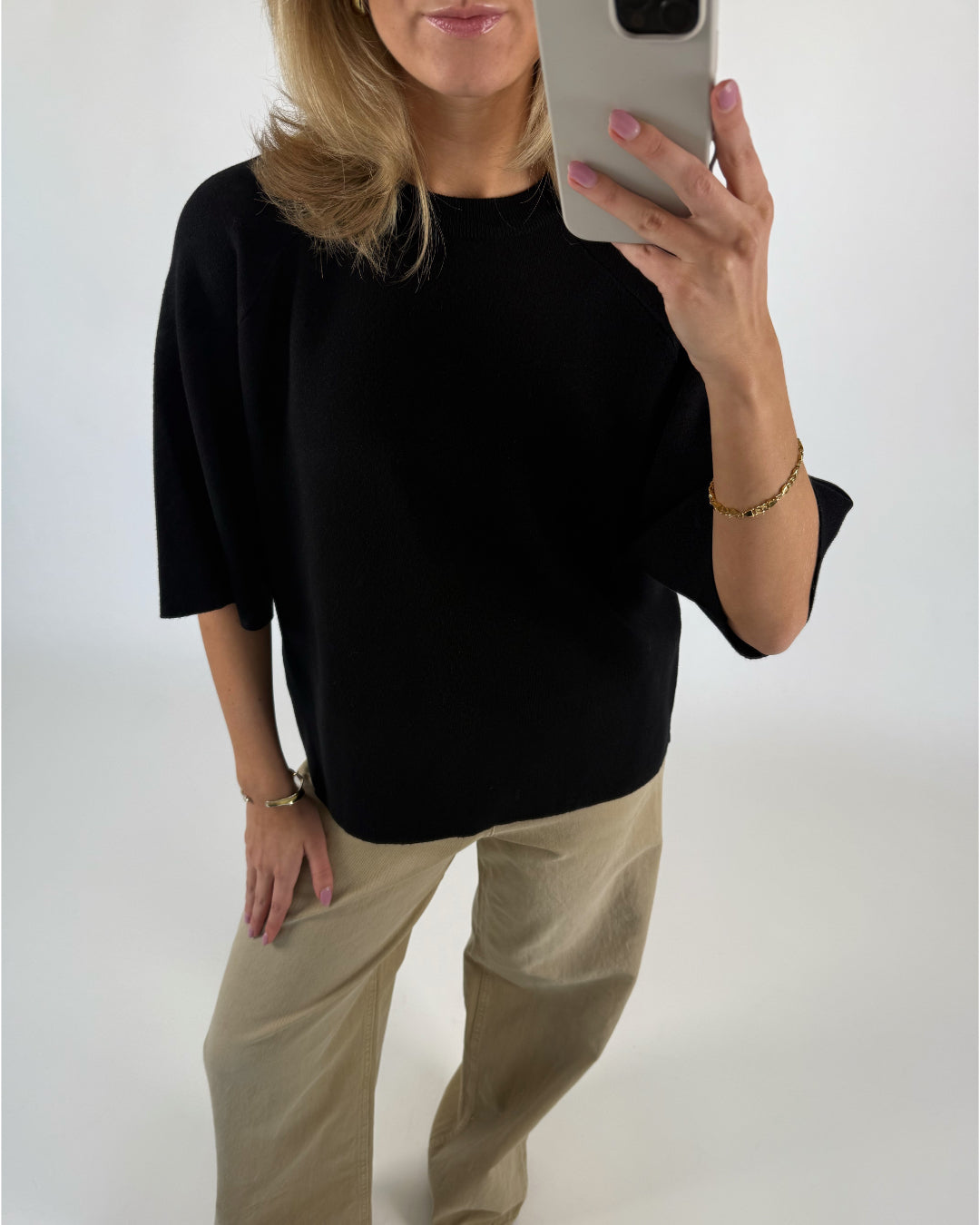 En person tager et selfiespejlbillede iført en sort trøje og beige bukser. De har en smartphone i hånden, der fremviser lyserød neglelak og et guldfarvet armbånd.