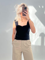 Person tager selfie med mobiltelefon, klædt i sort top og beige bukser. Håret er lyst og løst, i et rum med en grå væg og skygger.