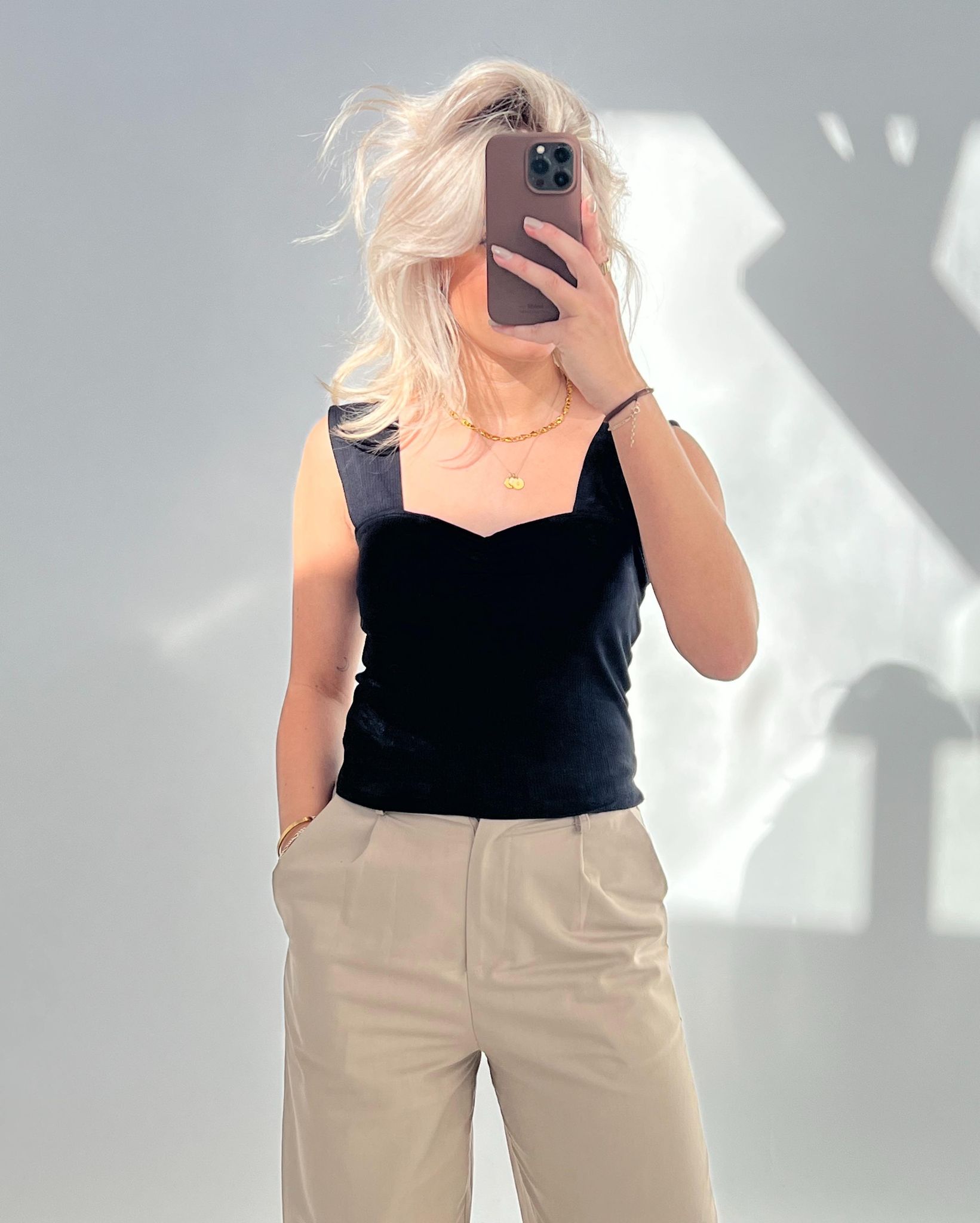 Person tager selfie med mobiltelefon, klædt i sort top og beige bukser. Håret er lyst og løst, i et rum med en grå væg og skygger.