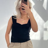 En person holder en smartphone op foran ansigtet i et rum med naturligt lys. Personen har blondt hår, sort top og beige bukser, og bærer guldsmykker.