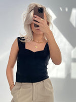 En person holder en smartphone op foran ansigtet i et rum med naturligt lys. Personen har blondt hår, sort top og beige bukser, og bærer guldsmykker.