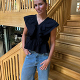 En person står afslappet med hænderne i lommerne, iført en sort bluse og jeans. Baggrunden er en indendørs trætrappe med moderne design.
