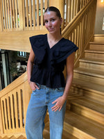 En person står afslappet med hænderne i lommerne, iført en sort bluse og jeans. Baggrunden er en indendørs trætrappe med moderne design.
