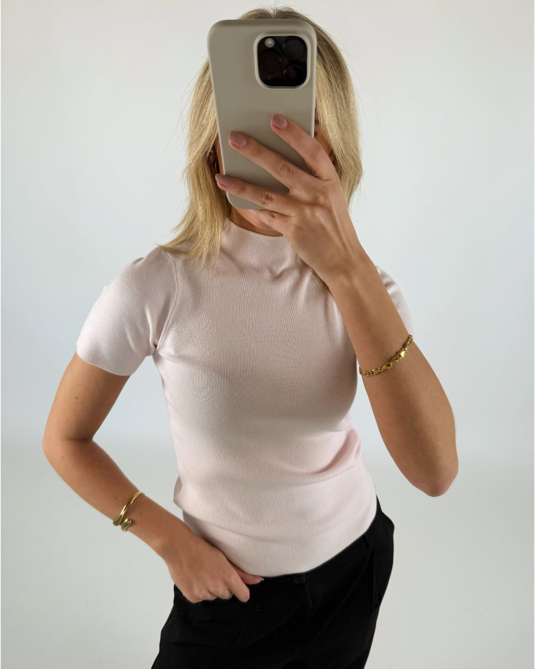 En person iført en lys t-shirt holder en smartphone foran ansigtet, i et hvidt, minimalistisk rum. Personen bærer guldsmykker på begge hænder.