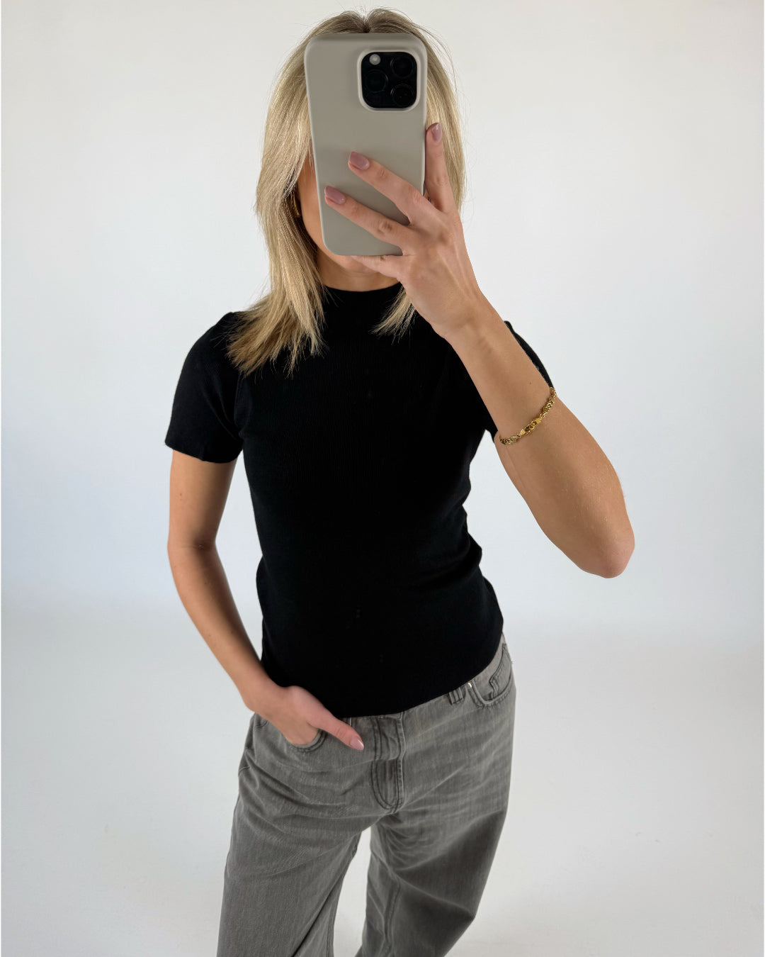 En person klædt i sort t-shirt og grå jeans tager et spejlbillede med en smartphone i en lys baggrund. En hånd hviler i bukselommen.