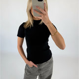 En person klædt i sort t-shirt og grå jeans tager et spejlbillede med en smartphone i en lys baggrund. En hånd hviler i bukselommen.