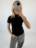 En person klædt i sort t-shirt og grå jeans tager et spejlbillede med en smartphone i en lys baggrund. En hånd hviler i bukselommen.
