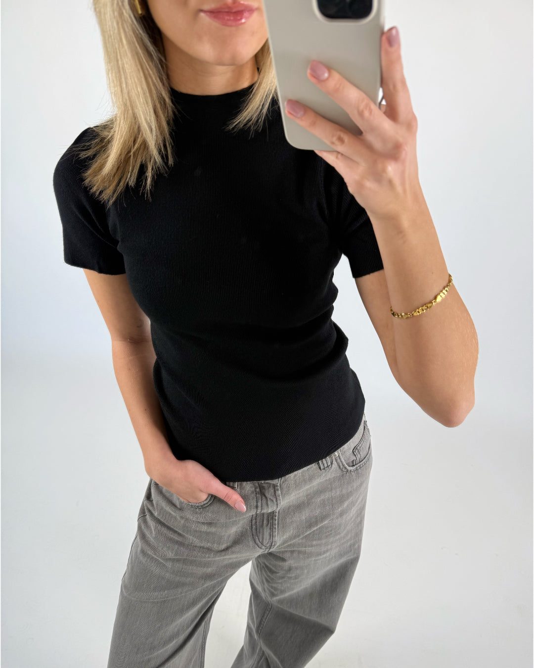En person i sort t-shirt tager en selfie med en mobiltelefon. Personen bærer grå bukser og et guldfarvet armbånd. Baggrunden er lys og neutral.