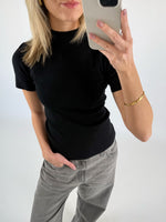 En person i sort t-shirt tager en selfie med en mobiltelefon. Personen bærer grå bukser og et guldfarvet armbånd. Baggrunden er lys og neutral.