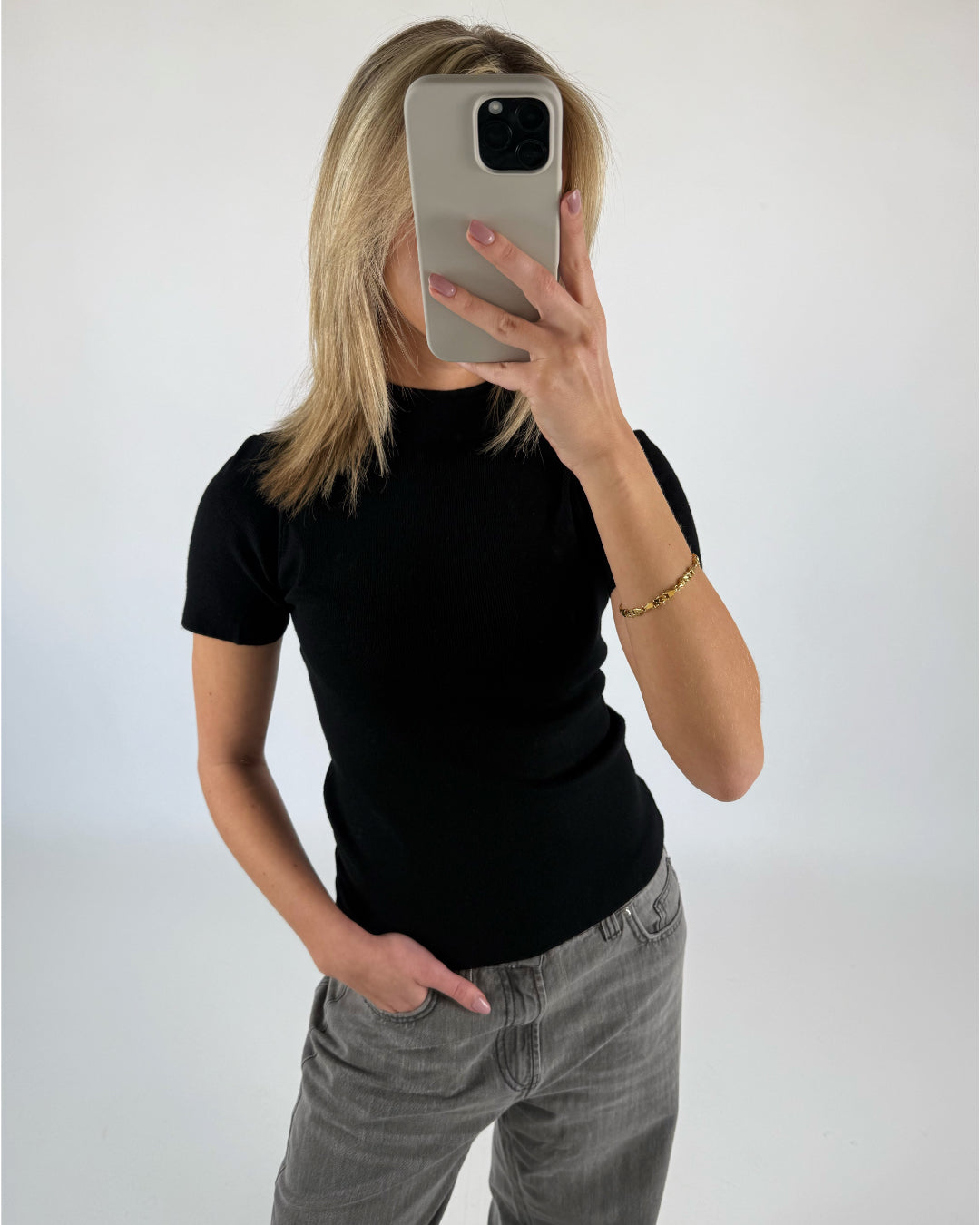 En person holder en smartphone foran ansigtet, står iført en sort trøje og grå jeans i et enkelt, hvidt rum.