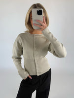 En person i en lys beige cardigan holder en smartphone op foran ansigtet. Personen står i et rum med en hvid baggrund og har en hånd i lommen på sine sorte bukser.