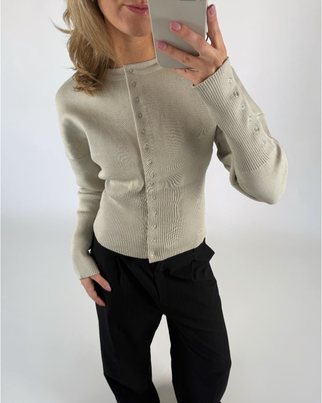 En person iført en lys sweater med knapper og sorte bukser tager et spejlbillede. Personen har løst hår og lakerede negle, i et neutralt oplyst rum.