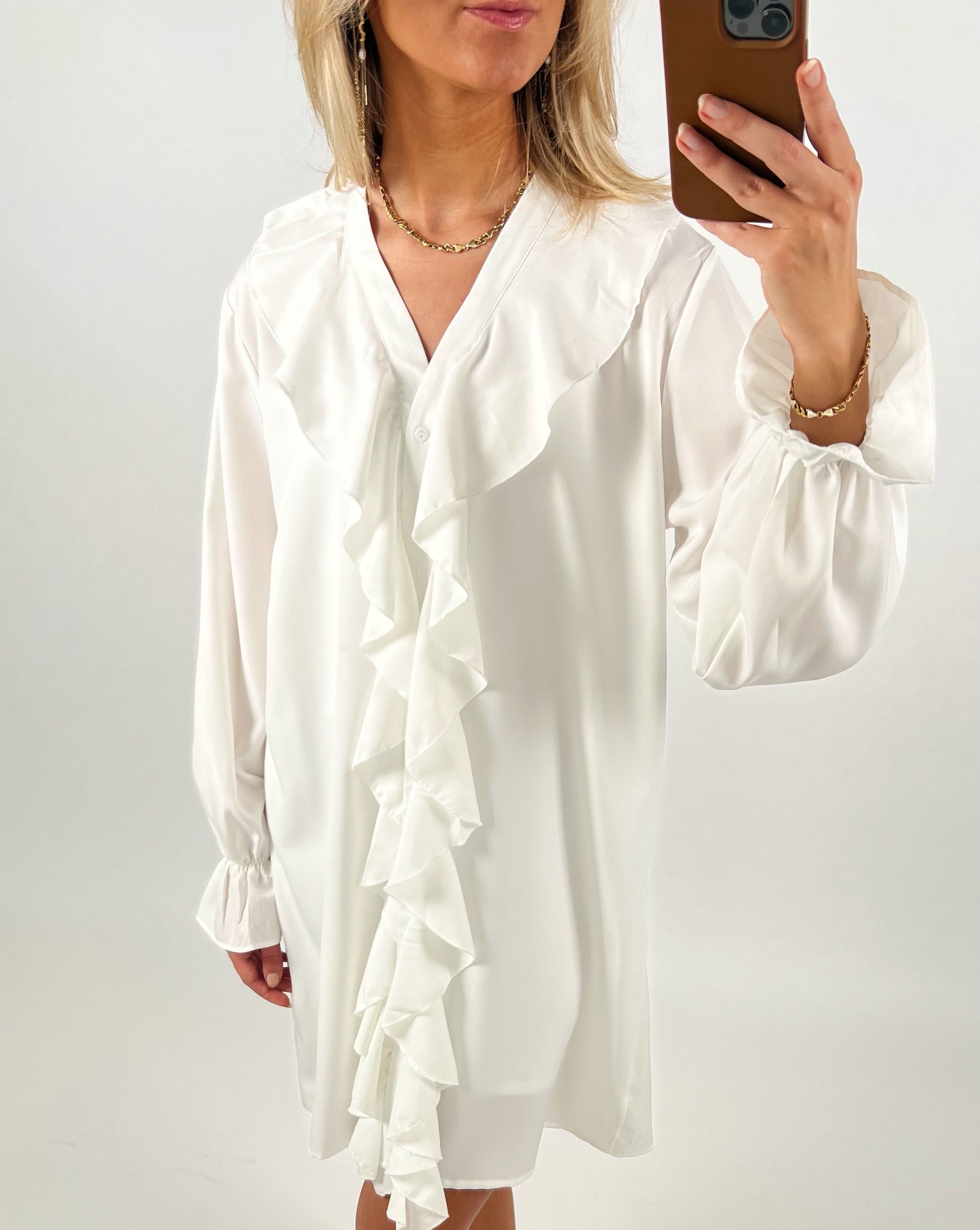 Kvinde iført hvid kjole med flæser bruger telefon til selfie. Hun står i et lyst rum, iført en kæde om halsen og et armbånd.