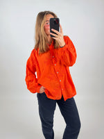 En person iført en orange skjorte tager et billede af sig selv med en smartphone. Personen står indendørs foran en neutral, lys baggrund.