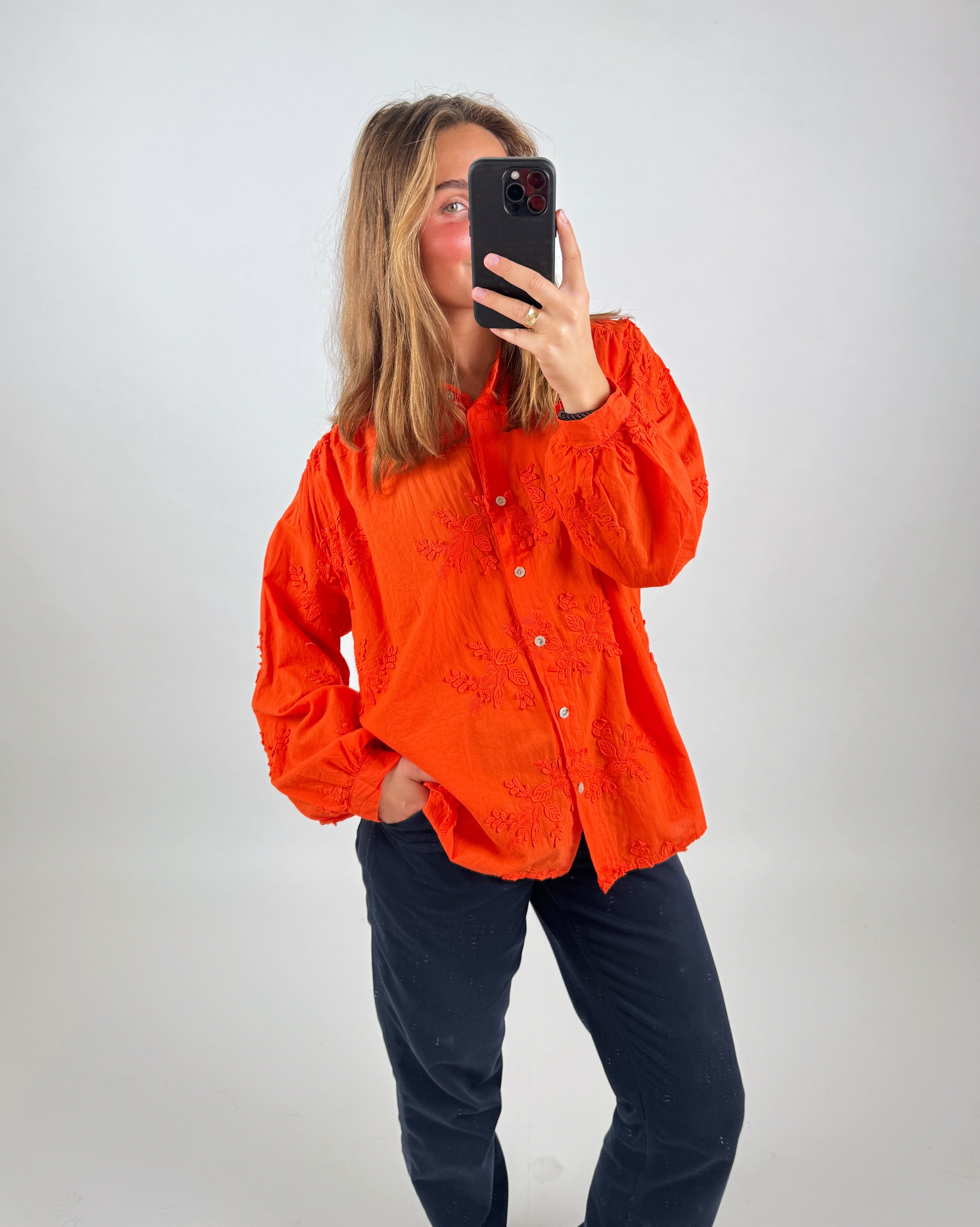En person iført en orange skjorte tager et billede af sig selv med en smartphone. Personen står indendørs foran en neutral, lys baggrund.