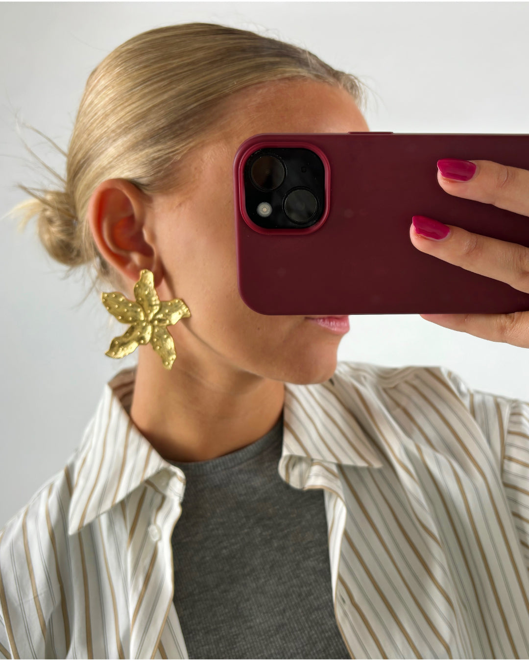 Person tager selfie i et spejl, iført en stor gylden blomsterørering. Personen har blondt hår i en knold, bærer en stribet skjorte over en grå bluse.