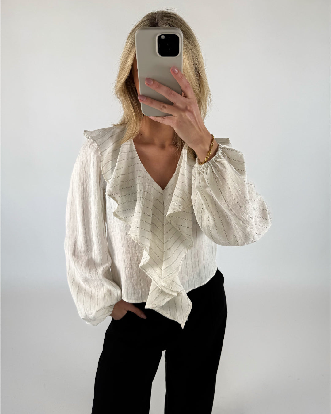 Person i en hvid bluse med flæser holder en mobiltelefon foran ansigtet. Personen står i et neutralt rum og har hånden i lommen på sorte bukser.