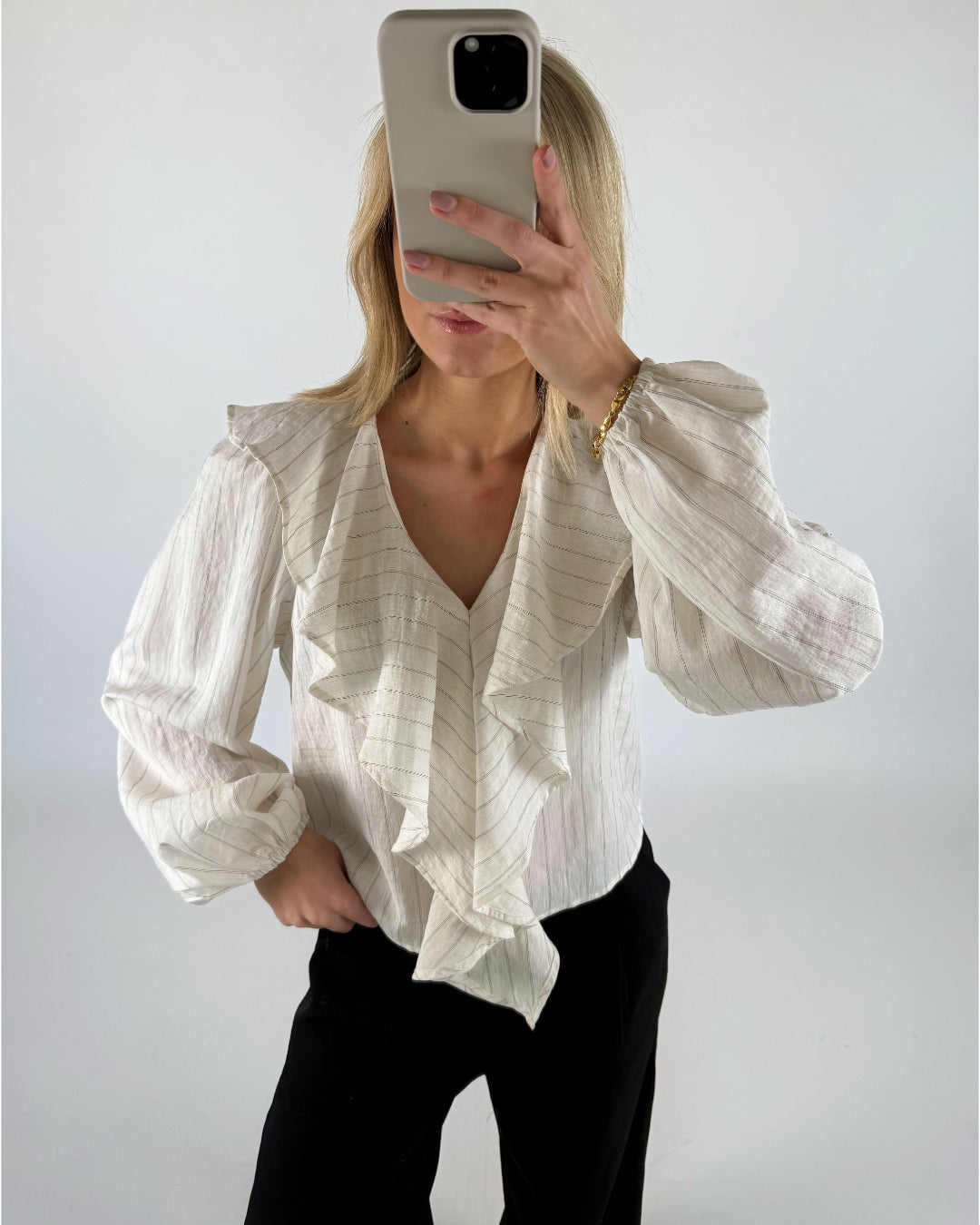En person i hvid bluse med flæser tager et spejlbillede med en smartphone. Baggrunden er en simpel, hvid væg.