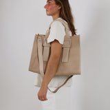 En kvinde bærer en stor beige taske på skulderen. Hun står i profil iført en hvid kjole mod en neutral, lysegrå baggrund.