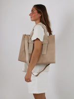 En kvinde bærer en stor beige taske på skulderen. Hun står i profil iført en hvid kjole mod en neutral, lysegrå baggrund.
