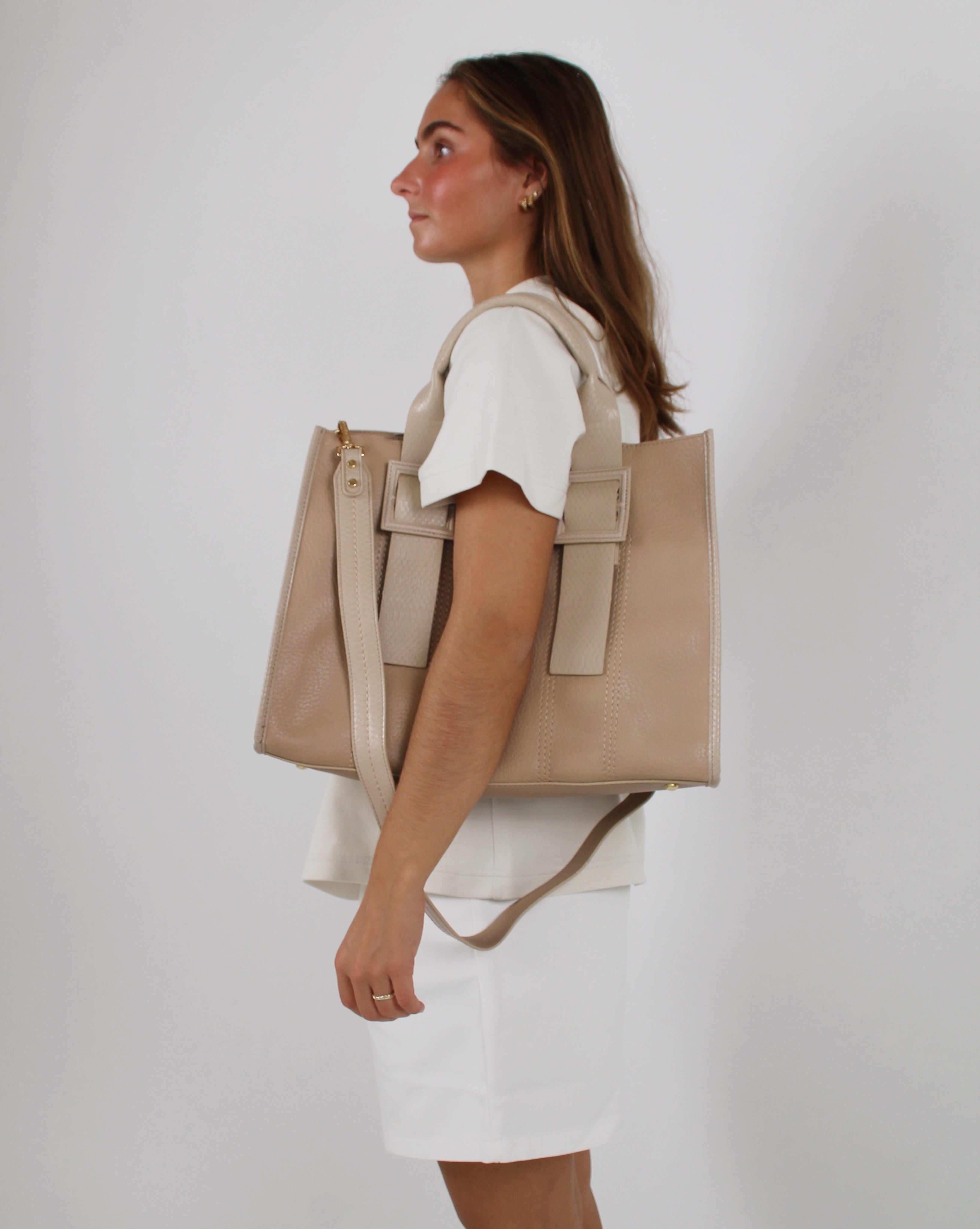 En kvinde bærer en stor beige taske på skulderen. Hun står i profil iført en hvid kjole mod en neutral, lysegrå baggrund.